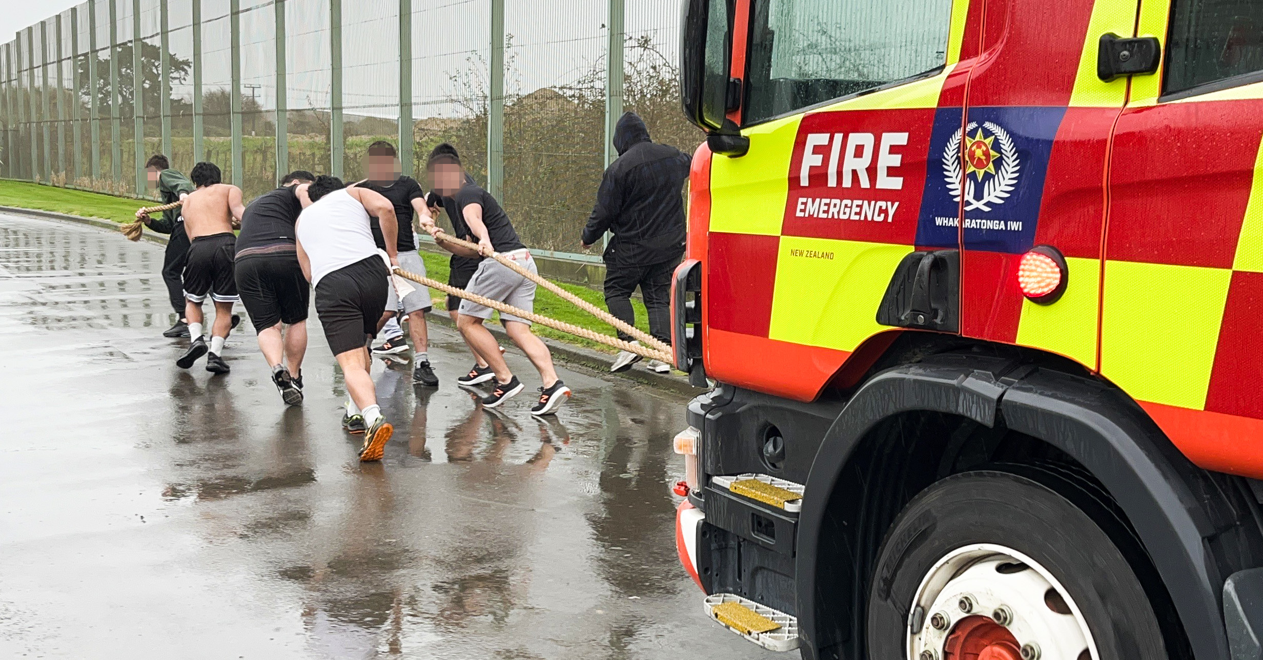A group of young people running forward holding two large ropes, pulling a fire truck behind them.